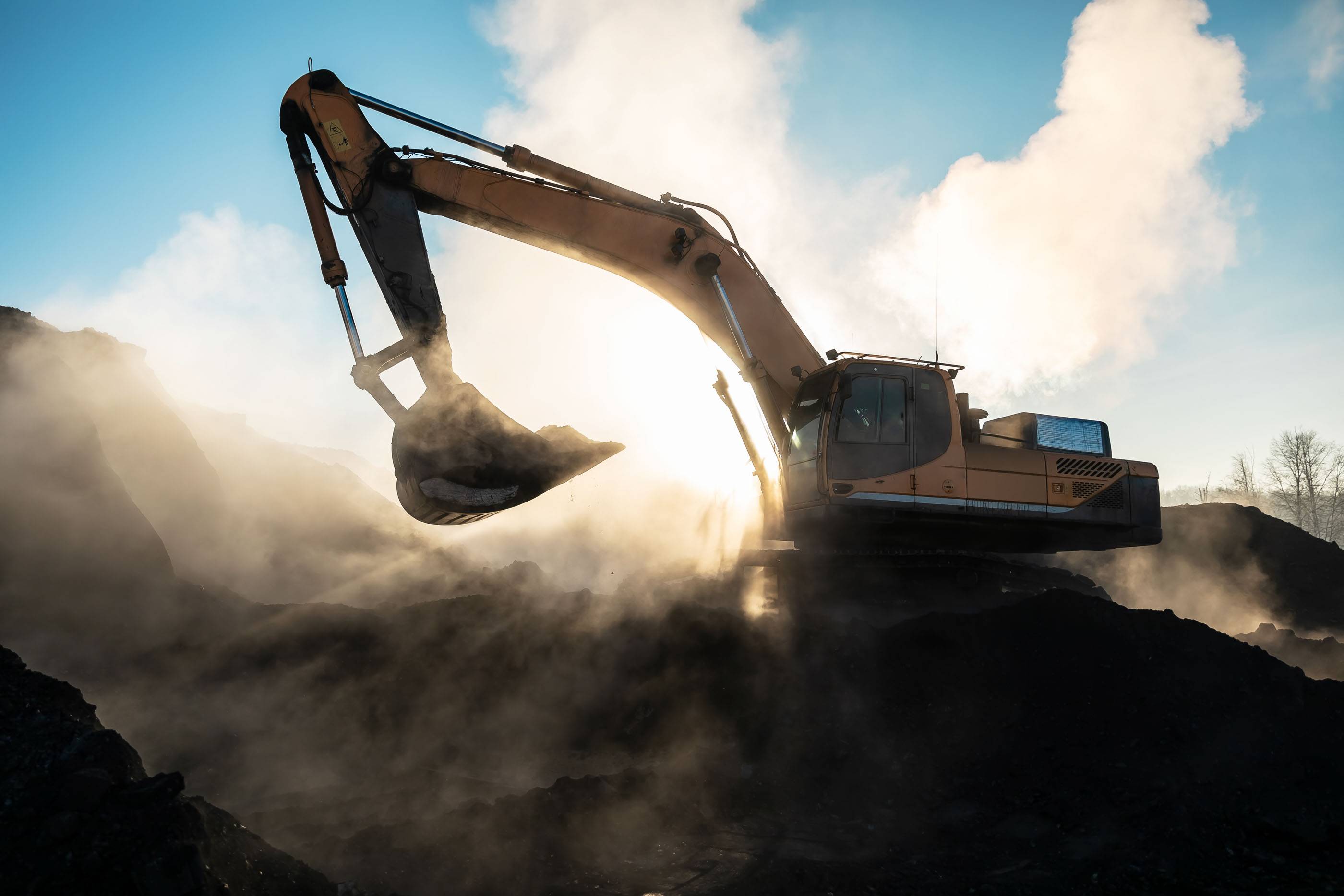 Большой желтый экскаватор в угольном разрезе загружает породу на фоне яркого солнца и чистого голубого неба. Техника на угольном разрезе. Карьерные машины и оборудование.