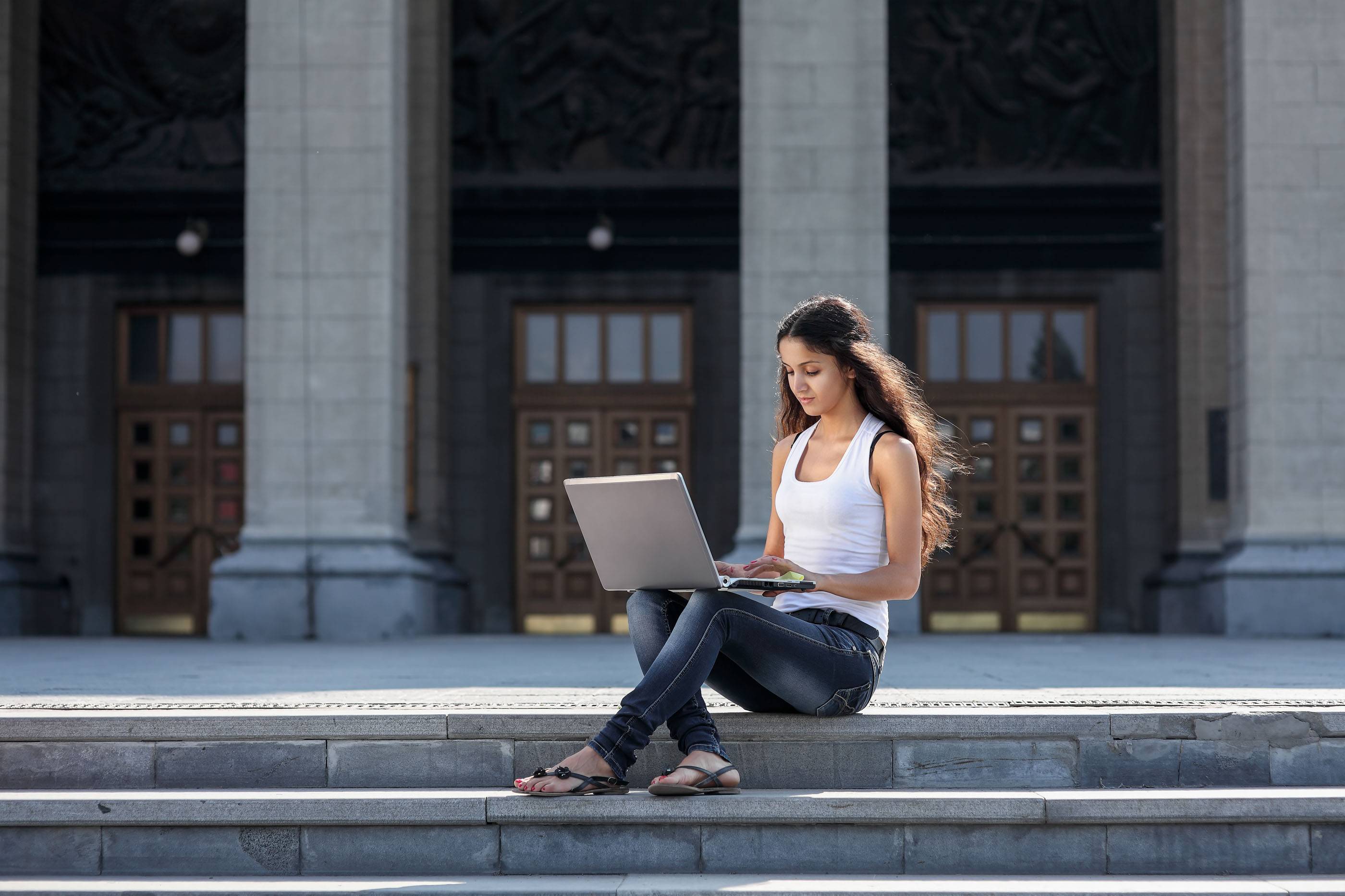 Молодая женщина с ноутбуком сидит на лестнице рядом с университетом