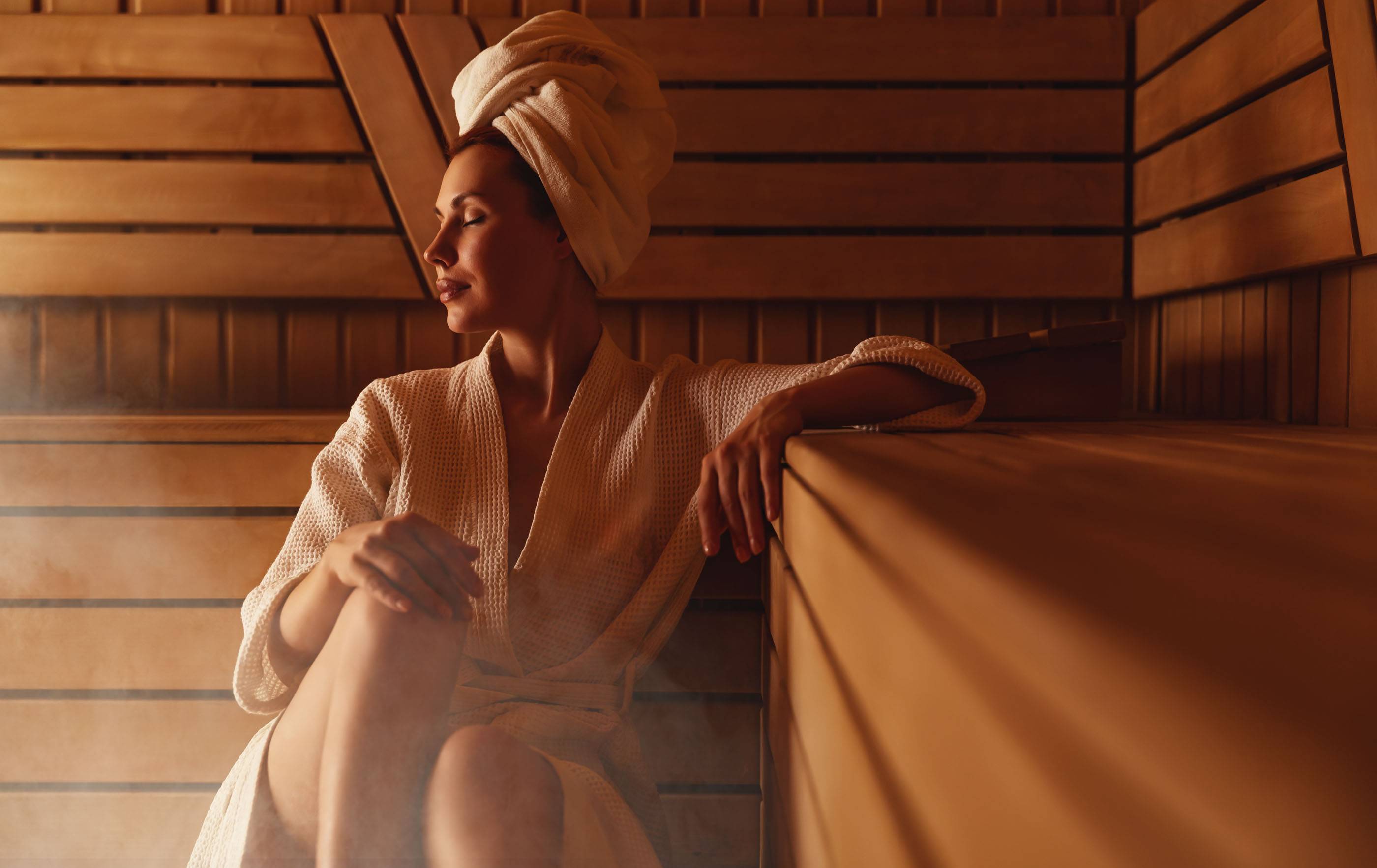 Молодая женщина отдыхает и потеет в горячей сауне, завернутая в полотенце. Девушка в сауне. Интерьер финской сауны, классическая деревянная парная с горячим паром. Русская баня. Отдых в горячей бане с паром.