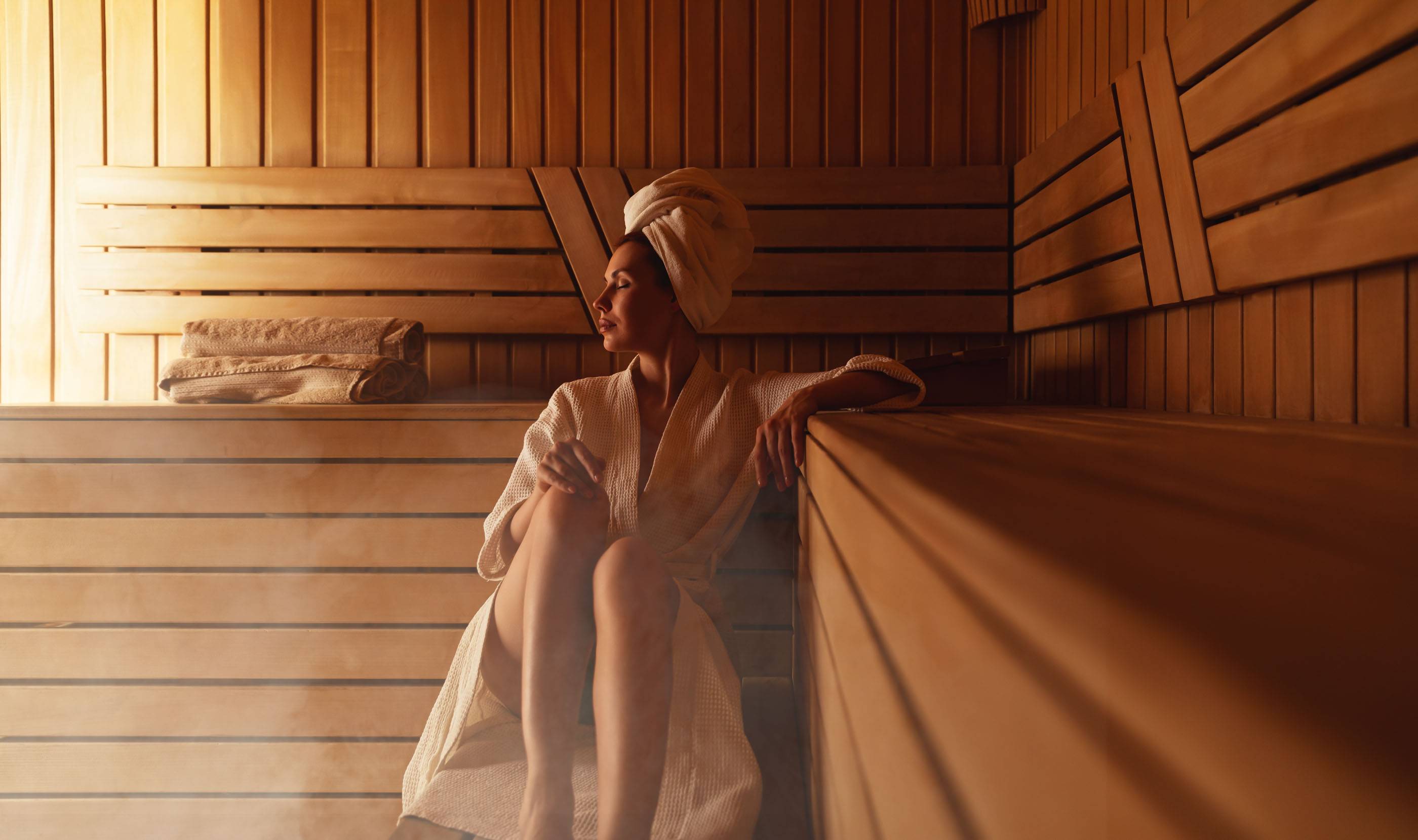 Молодая женщина отдыхает и потеет в горячей сауне, завернутая в полотенце. Девушка в сауне. Интерьер финской сауны, классическая деревянная парная с горячим паром. Русская баня. Отдых в горячей бане с паром.