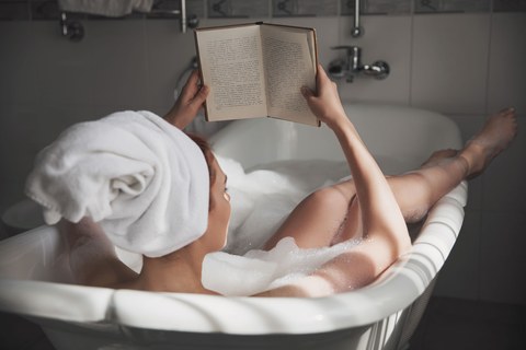 Вид сзади: красивая молодая женщина читает книгу, наслаждаясь ванной с пеной. Дама в отпуске в отеле лежит в расслабляющей ванне с бумажной книгой. Молодая женщина читает книгу, отдыхая в ванне.