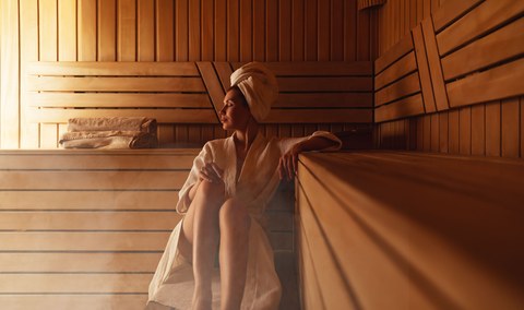 Молодая женщина отдыхает и потеет в горячей сауне, завернутая в полотенце. Девушка в сауне. Интерьер финской сауны, классическая деревянная парная с горячим паром. Русская баня. Отдых в горячей бане с паром.
